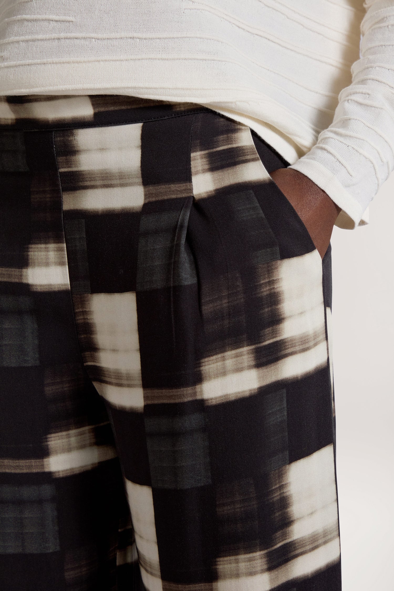 Close-up of a person donning a white textured top and Blurred Check Trouser, featuring an elasticated waistband in black and white. The person's hand rests in the pant pocket.