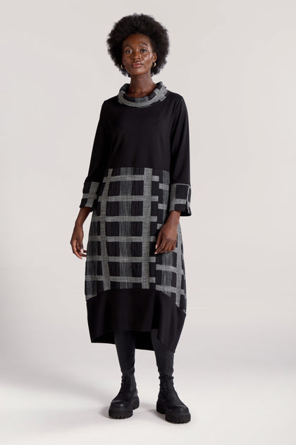 A woman with an afro hairstyle poses slightly off camera while wearing a Checked Cowl Neck Dress, characterized by a black long-sleeve top and a gray check print on the bottom half and sleeves; she completes her outfit with black boots.