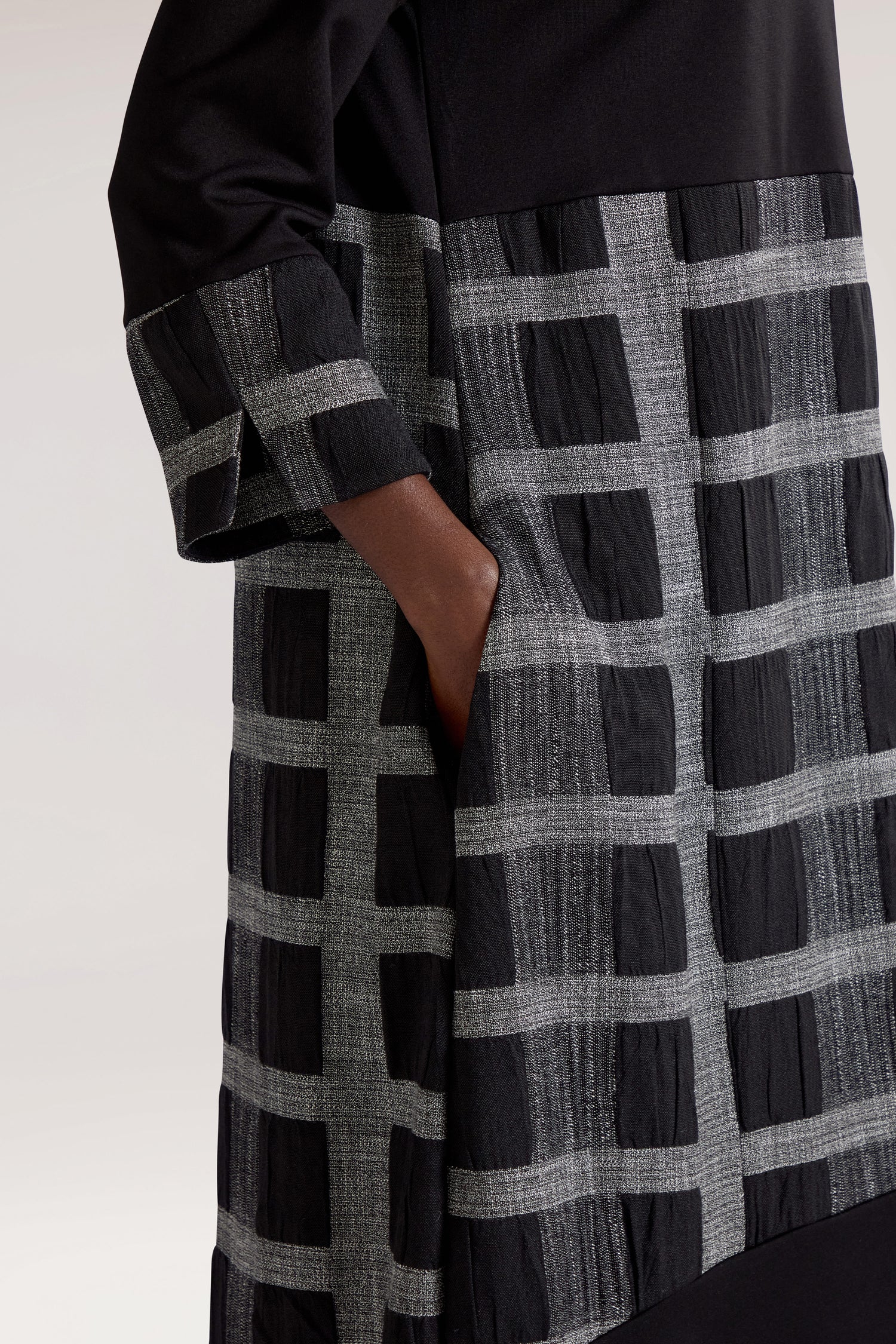 Close-up of an individual donning the Checked Cowl Neck Dress, showcasing a long-sleeved black outfit adorned with a grey grid pattern, with one hand tucked into a side pocket.