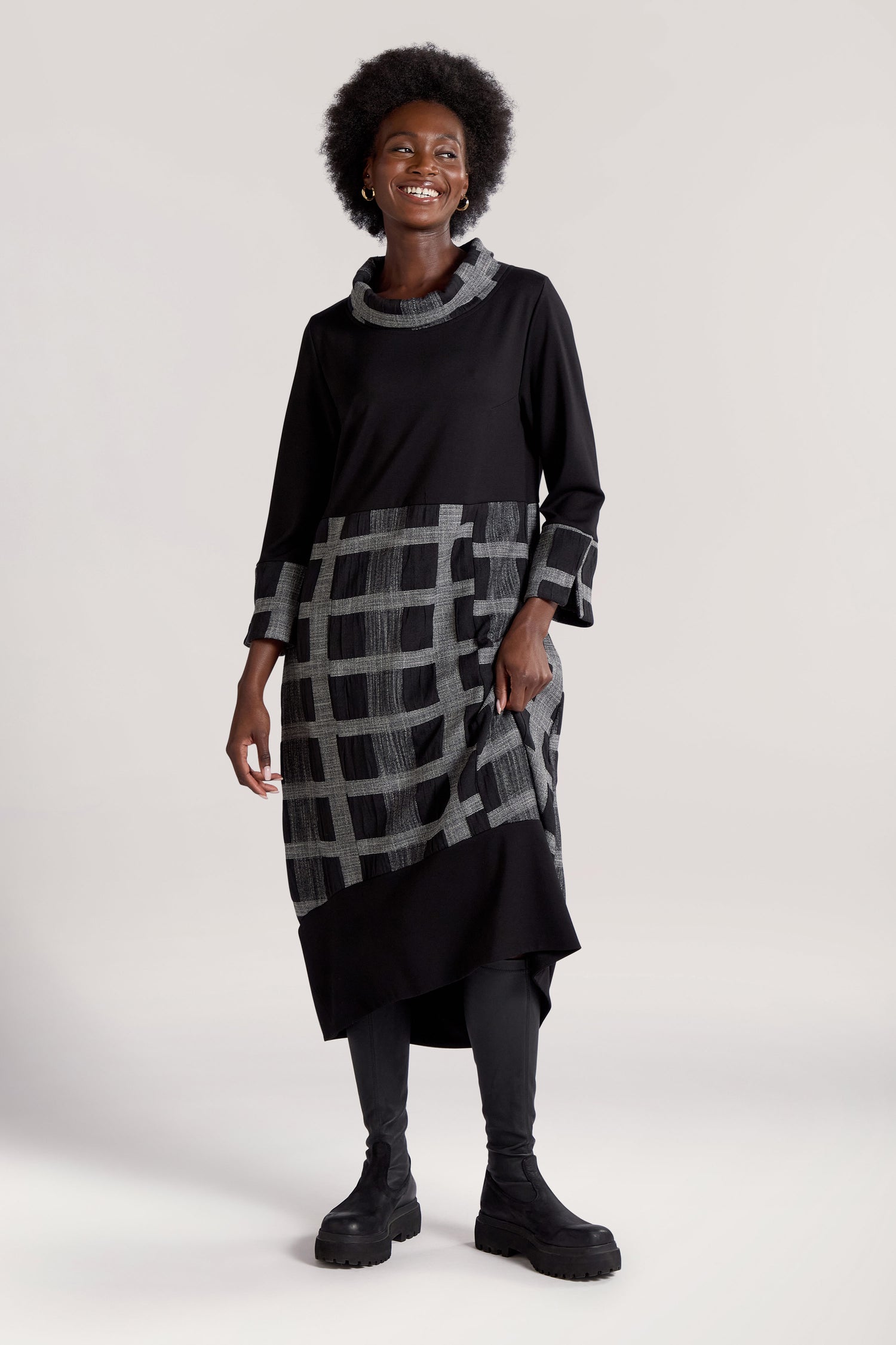 A person stands smiling, wearing the Checked Cowl Neck Dress in a black and grey check print, paired with black leggings and boots. The dress features a bubble-style skirt that adds a playful touch. They have short, curly hair. The background is a plain, light-colored backdrop.