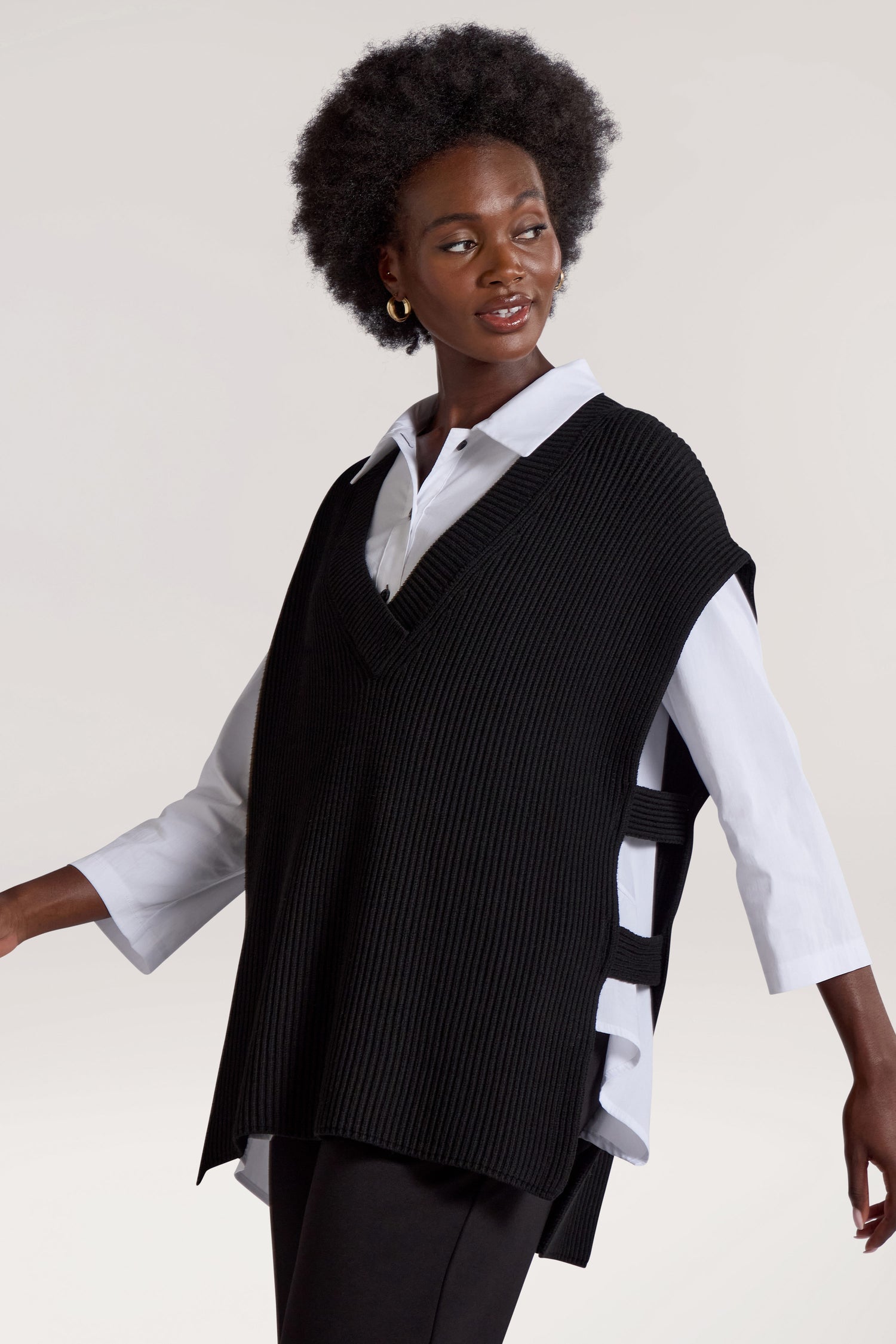 A woman with an afro hairstyle wears a white blouse under a black, sleeveless, ribbed V Neck Tabard with side ties. She is facing slightly to the side and looking away from the camera.