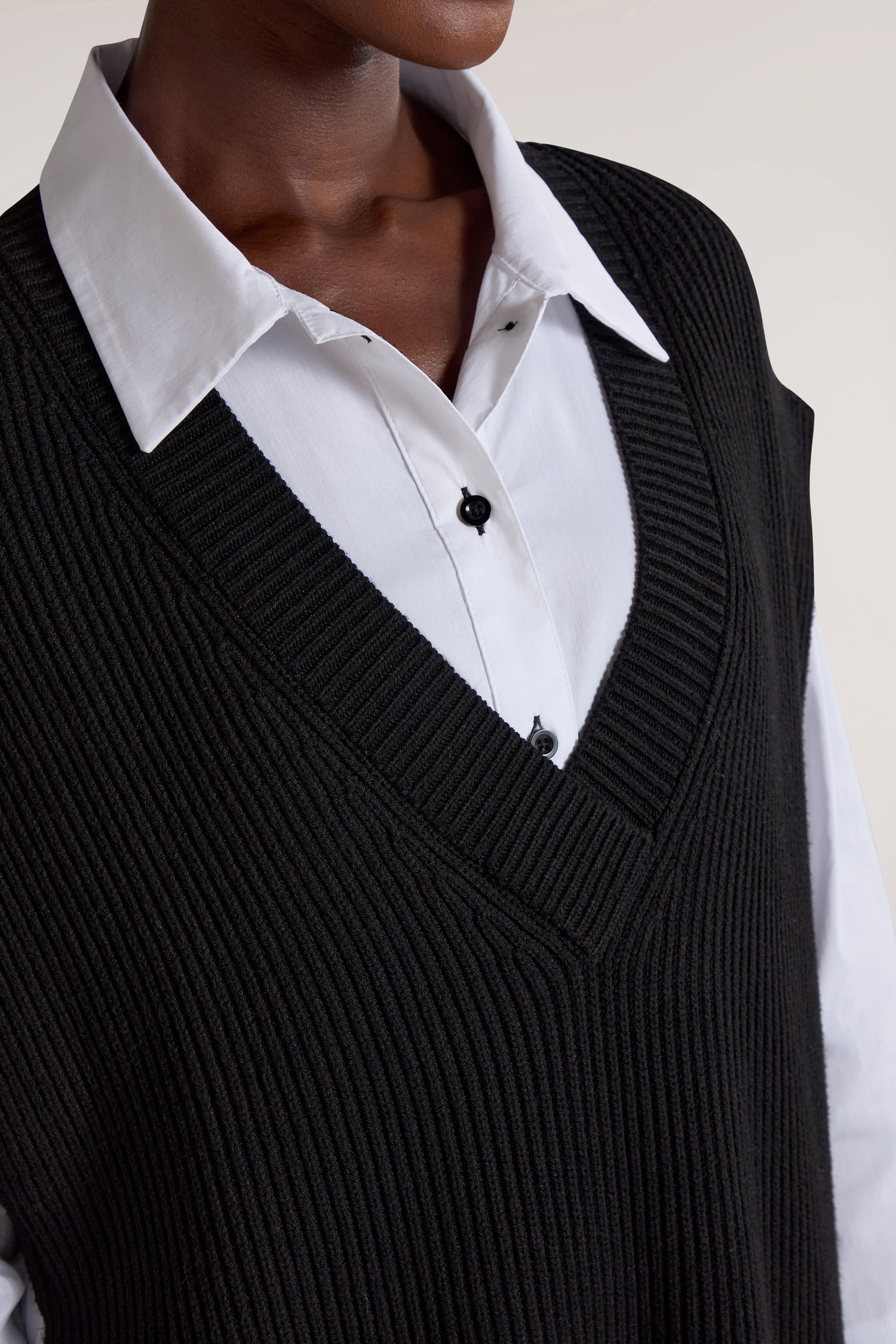 Close-up of a person wearing a white collared shirt underneath the versatile V Neck Tabard in black, making it a perfect layering piece.