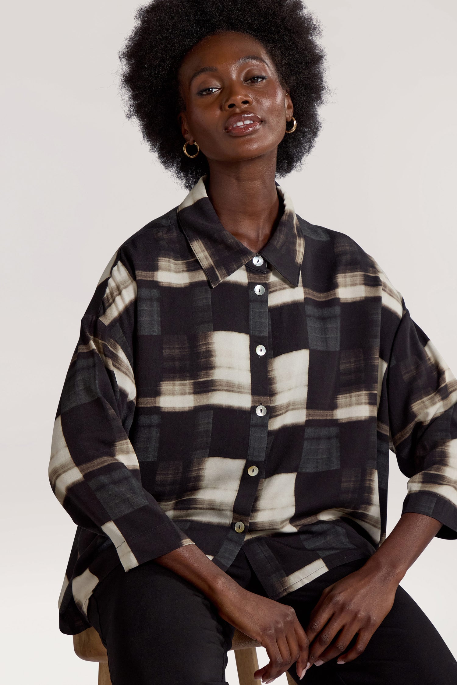 A person with an afro hairstyle, wearing the Blurred Check Boxy Shirt, sits on a stool against a plain background.