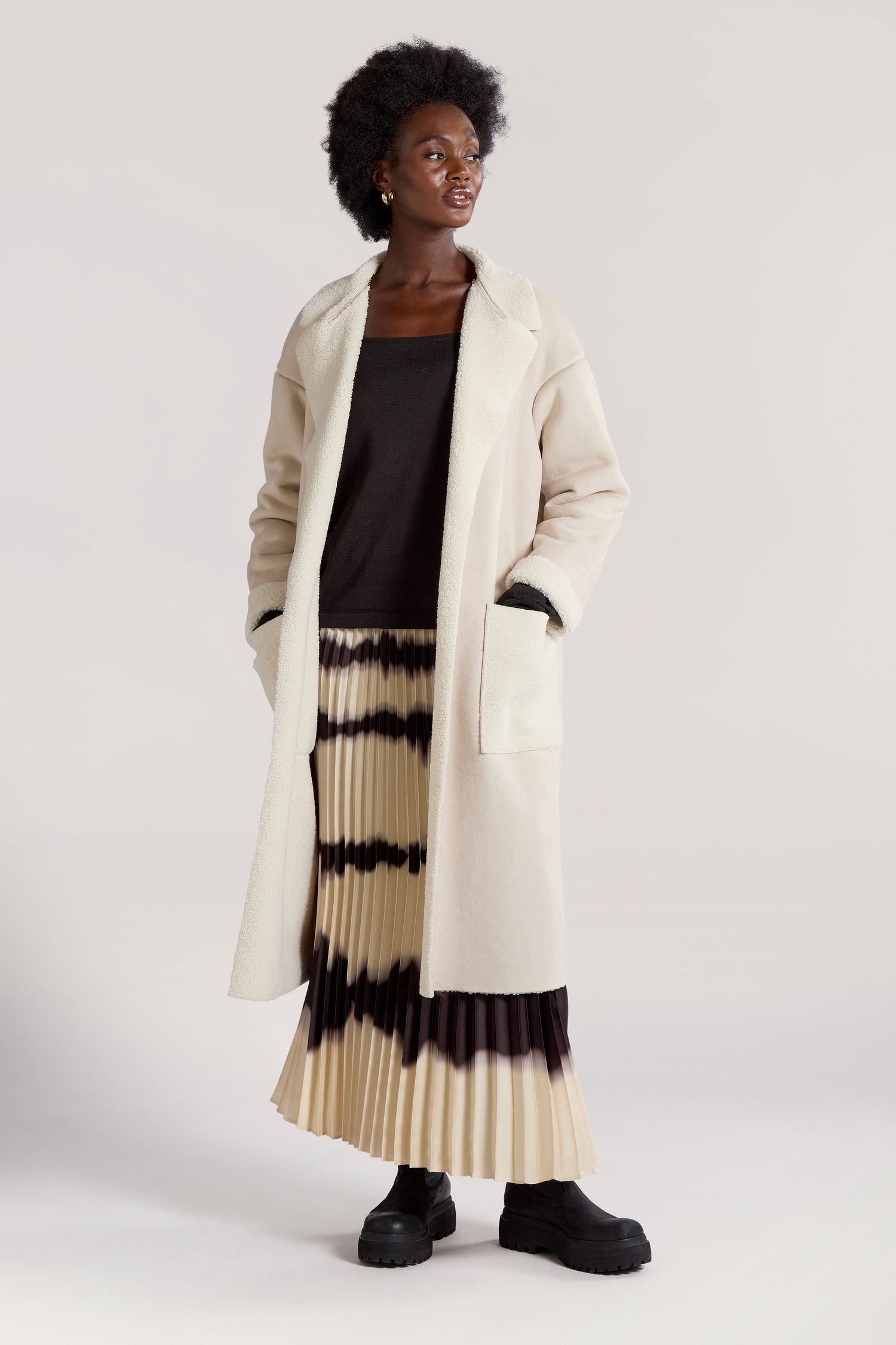 A person standing in a long, cream-colored Faux Sheepskin Coat over a black top and a cream and black pleated skirt, wearing black boots. Hands are in the coat pockets, and they are looking slightly to the side with a modern minimalist vibe.