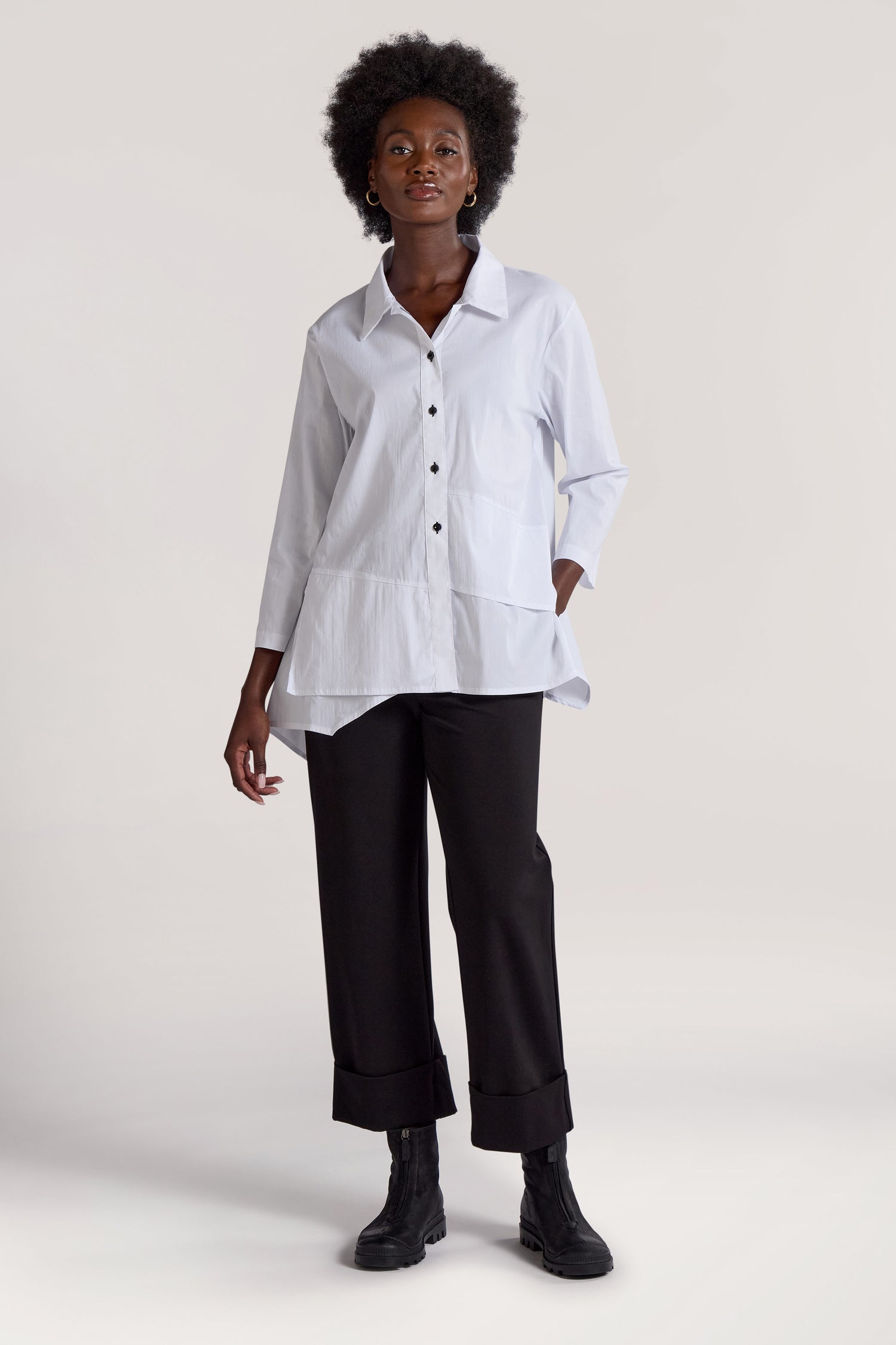 A person is standing and looking forward, wearing a white long-sleeve blouse, black Ponte Cuff Hem Trousers with an elasticated waistband, and black boots. With one hand in their pocket, they pose against a plain background, embodying a minimalist approach to fashion.