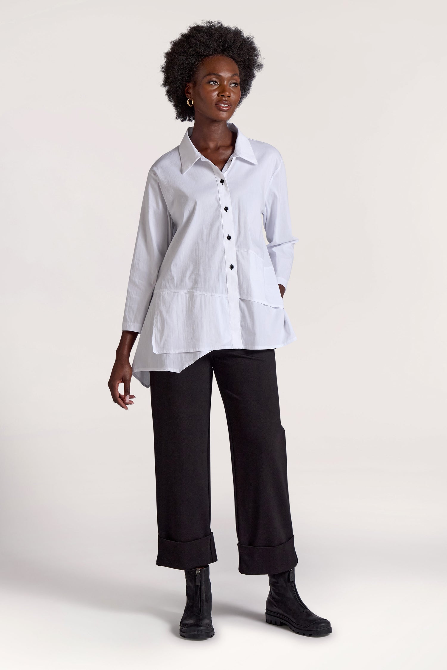 Against a plain background, a person with short curly hair embodies a minimalist approach. They wear a loose white button-up shirt and black Ponte Cuff Hem Trousers with rolled cuffs, paired with black ankle boots.