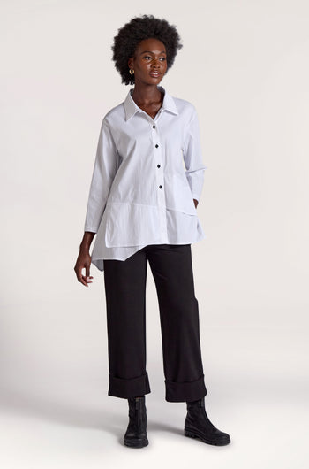 Against a plain background, a person with short curly hair embodies a minimalist approach. They wear a loose white button-up shirt and black Ponte Cuff Hem Trousers with rolled cuffs, paired with black ankle boots.