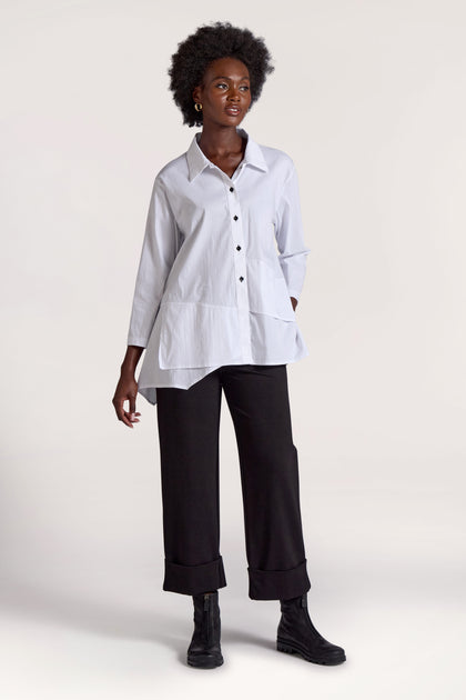 Against a plain background, a person with short curly hair embodies a minimalist approach. They wear a loose white button-up shirt and black Ponte Cuff Hem Trousers with rolled cuffs, paired with black ankle boots.