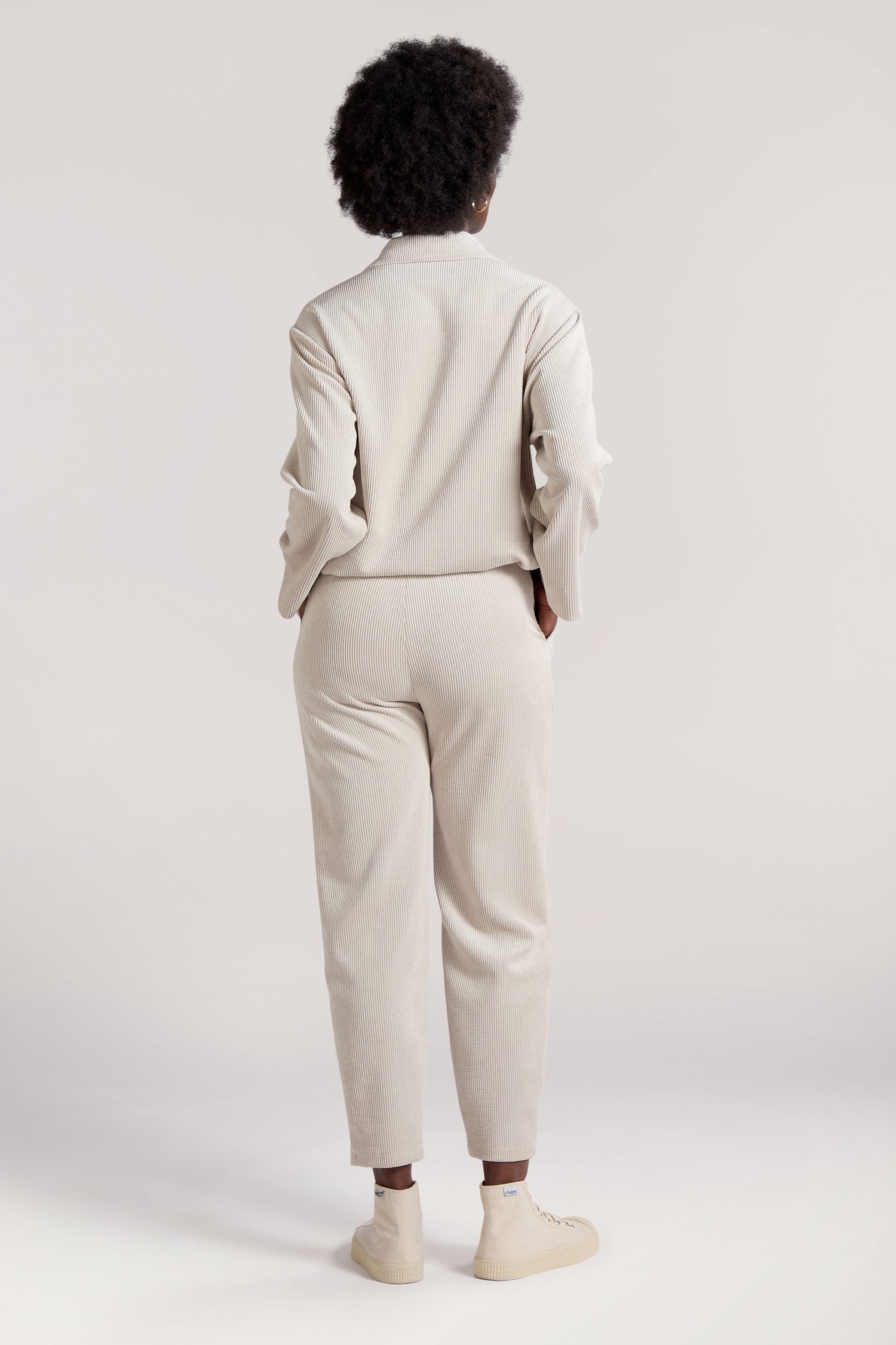 A person with short curly hair is standing with their back to the camera, wearing light beige Cord Trousers made from premium corduroy fabric and cream-colored shoes.