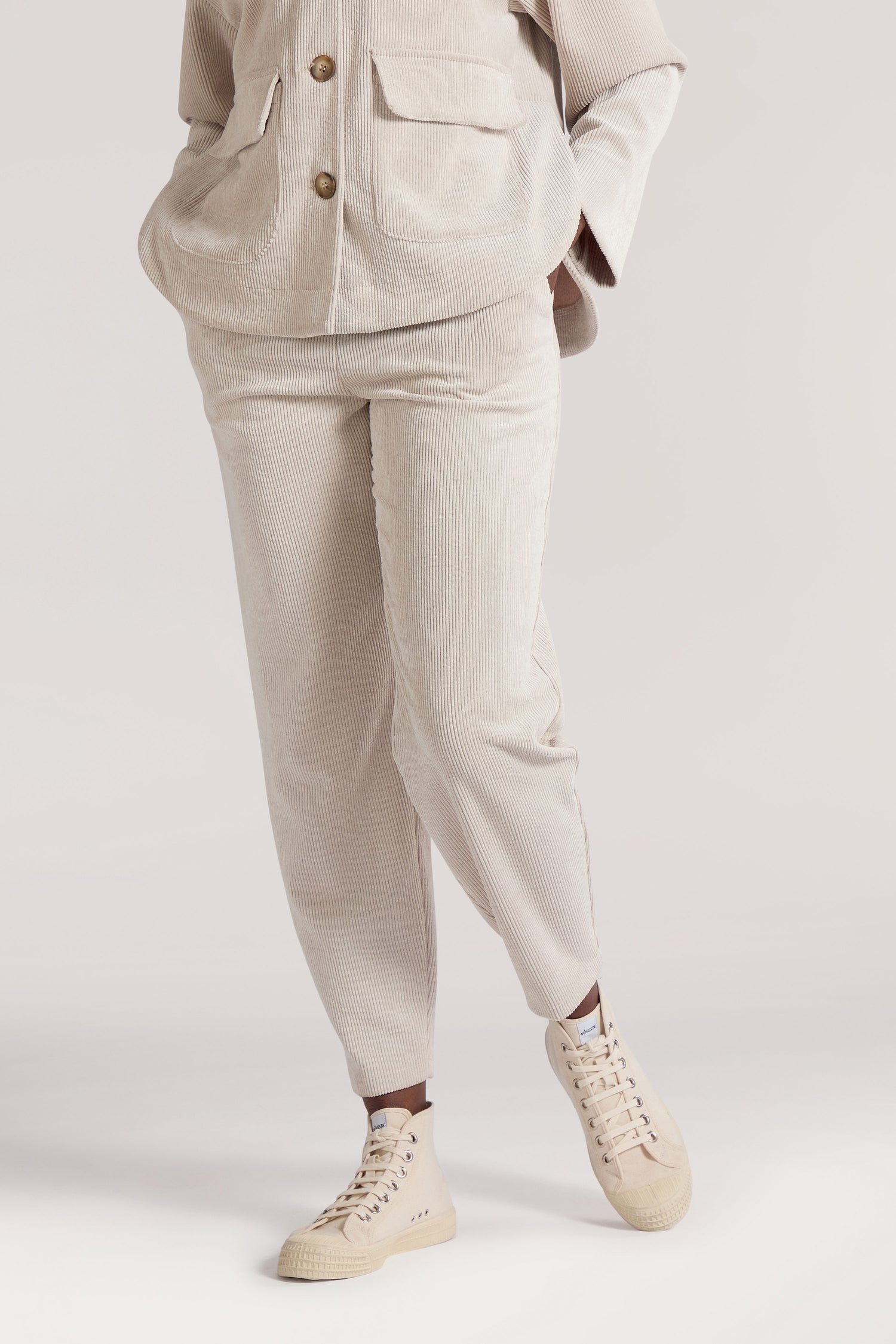 A person stands with hands in pockets, wearing a beige jacket and **Cord Trouser** made from premium corduroy fabric, and off-white sneakers.