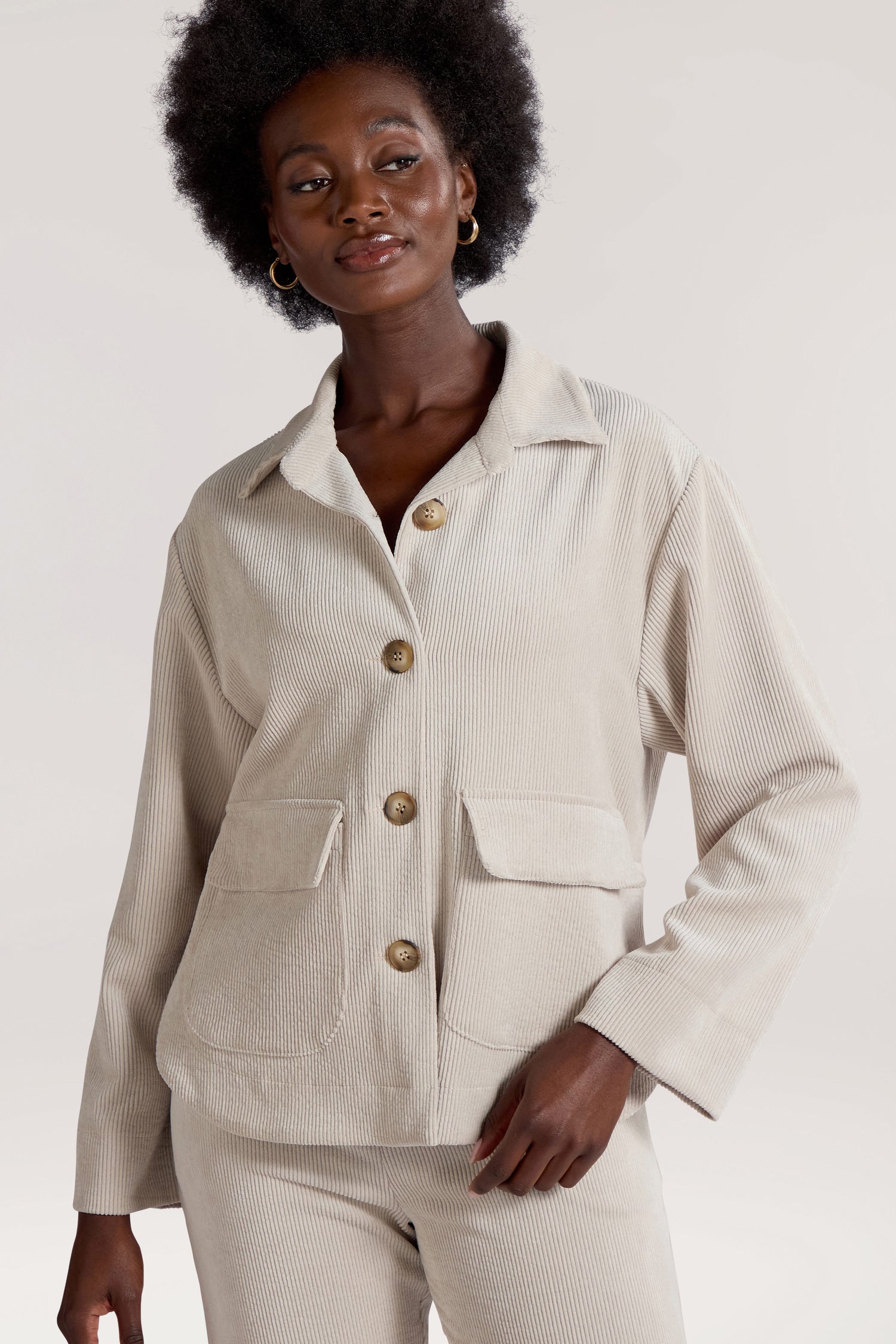 A person with an afro hairstyle is wearing a light-colored, long-sleeve **Cord Jacket** featuring front pockets, crafted from corduroy fabric. They are smiling slightly and looking to the side, exuding a minimalist appeal.