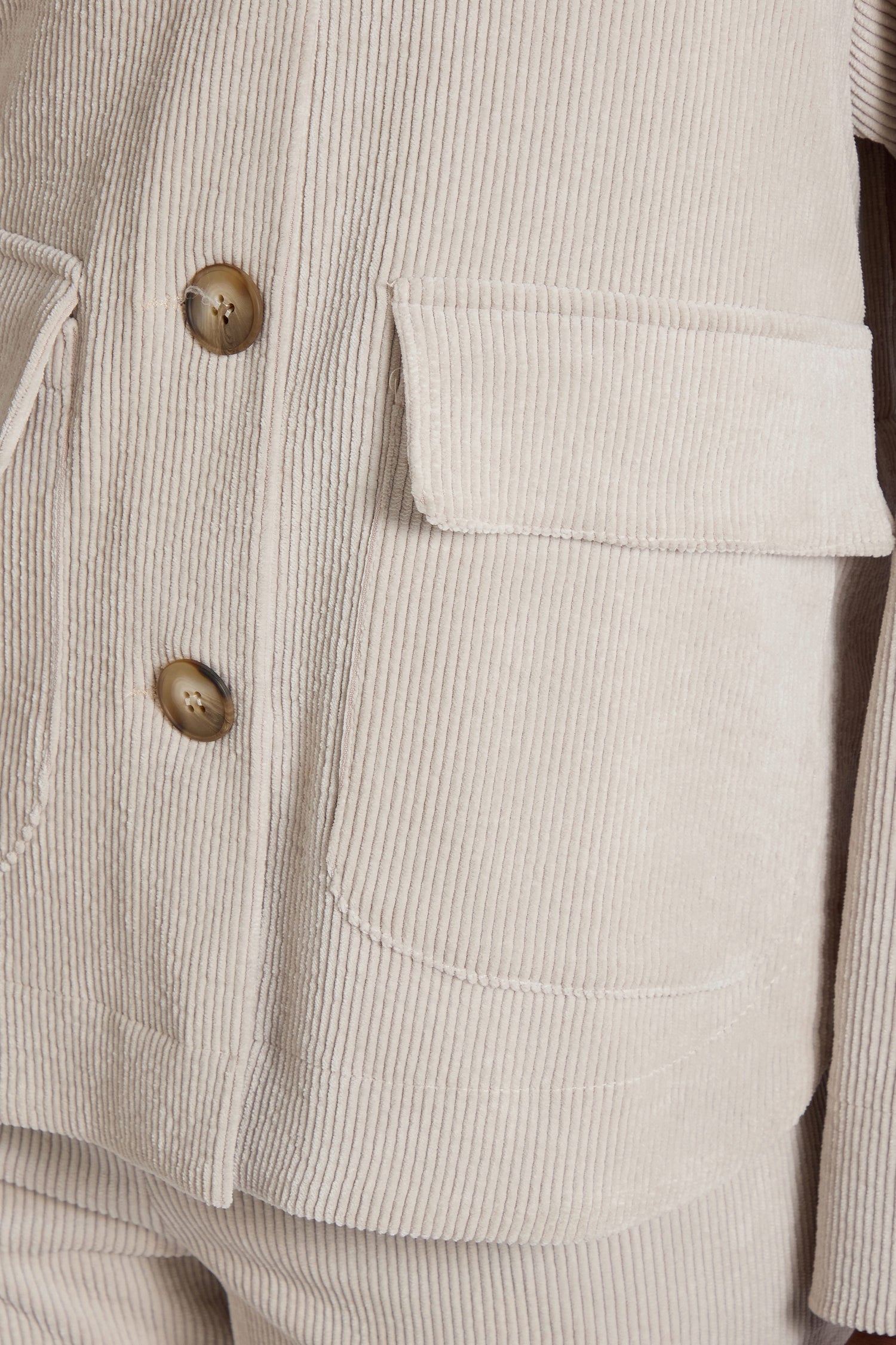 Close-up of the Cord Jacket in beige, featuring two brown buttons and a large pocket on the left side, exuding a minimalist appeal. The texture and vertical ridges of the corduroy fabric are clearly visible.