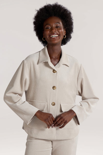 A person stands smiling while wearing the Cord Jacket in light beige, featuring pockets and exuding a minimalist appeal.