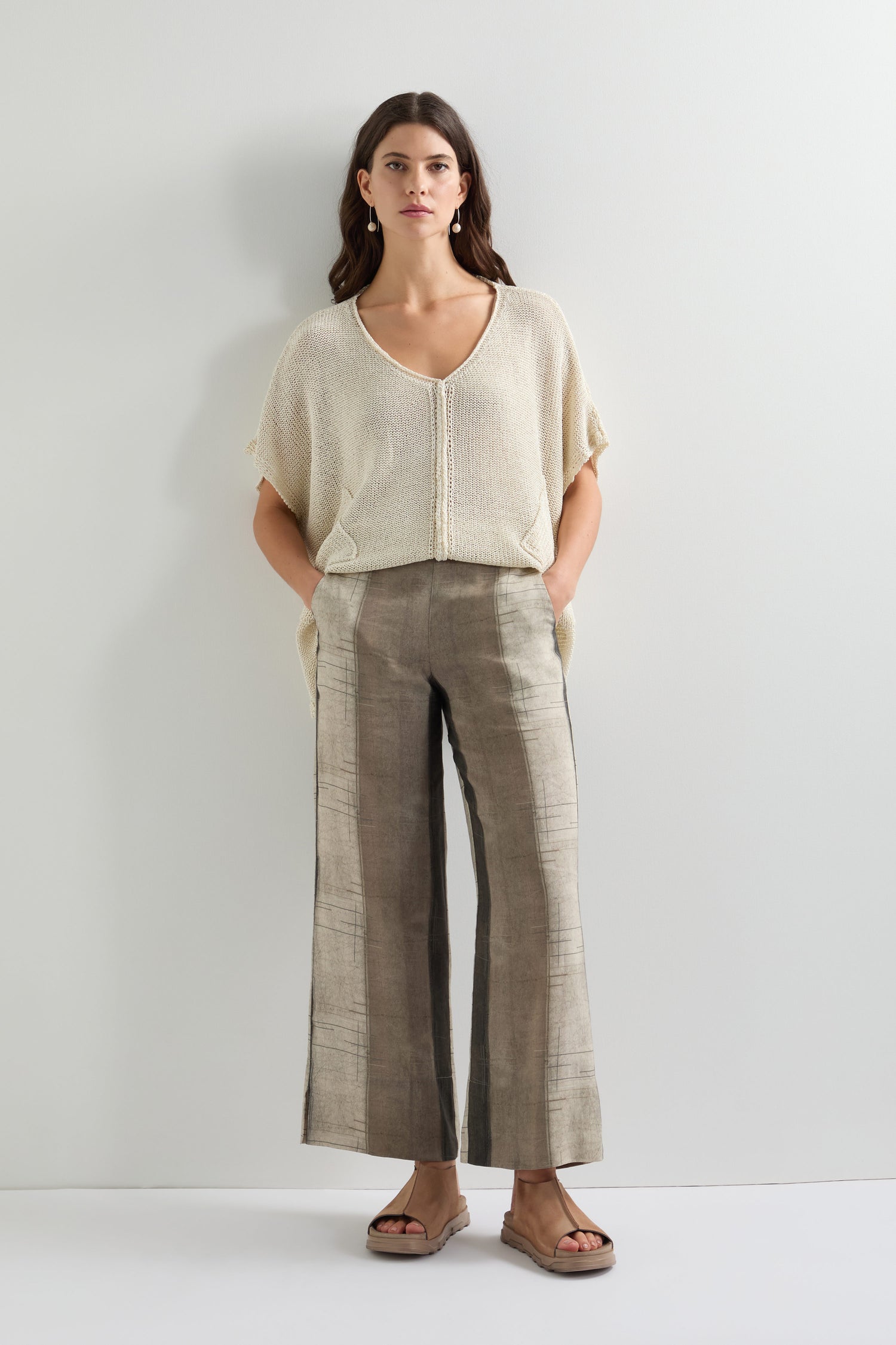 A woman wearing the Wide Stripe Trouser, a lightweight and breathable gray wide-leg pant, paired with a beige knitted top, stands against a white background with her hands in her pockets.