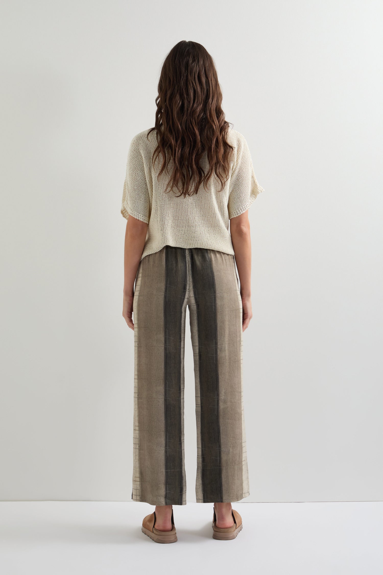 A person with long hair faces away, styled in a cream knit top and Wide Stripe Trouser featuring a relaxed wide-leg silhouette. Paired with sandals against a plain backdrop, this ensemble showcases lightweight breathable fabric for effortless elegance.