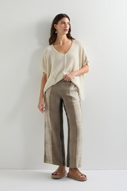 Against a plain wall, a person wears a loose beige top and Wide Stripe Trouser with a relaxed wide-leg silhouette. Paired with brown sandals, their long hair gently cascades as they gaze to the side.