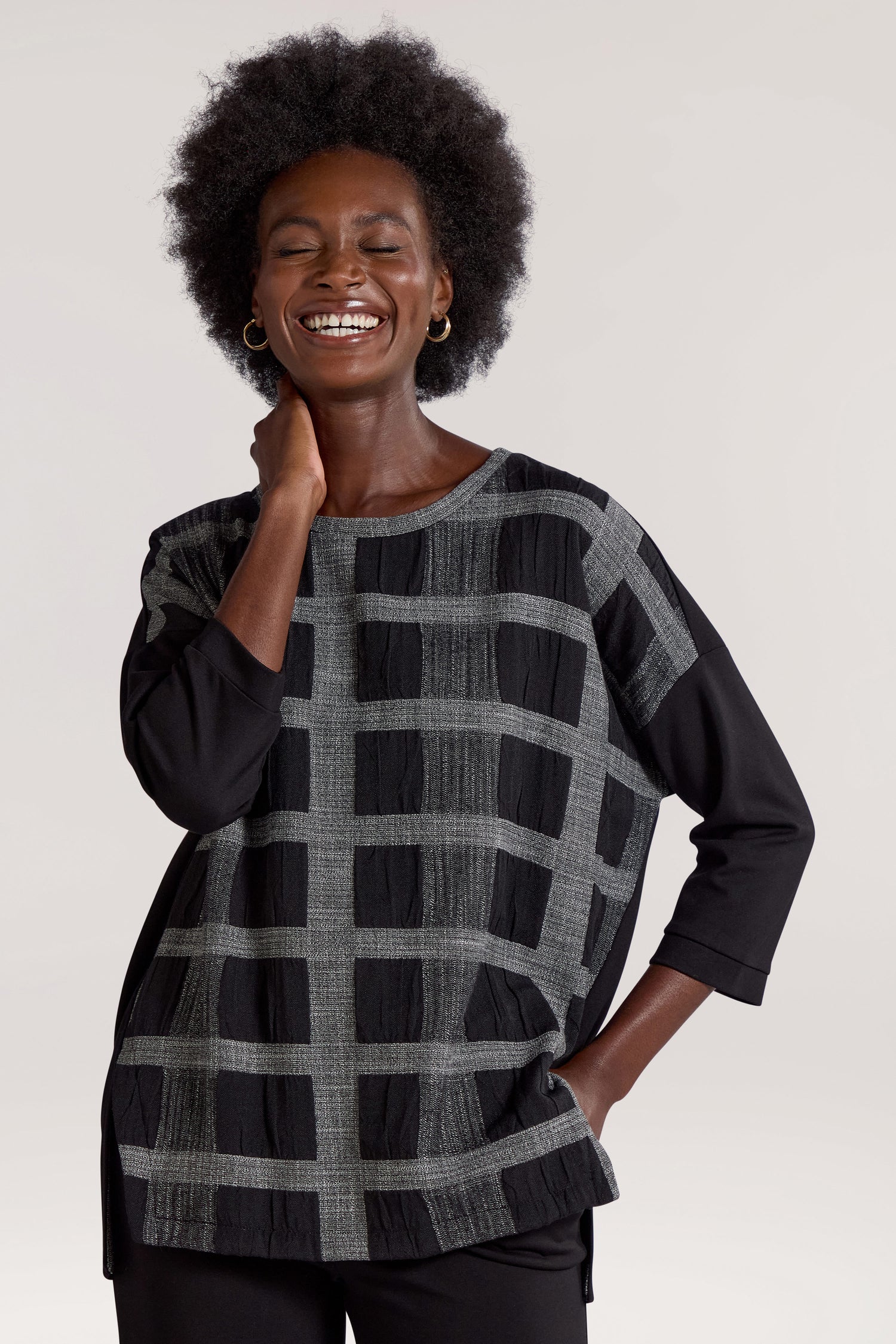 A person with short curly hair smiles, wearing the Checked Tunic in black and gray with three-quarter sleeves and black pants, standing with one hand in their pocket, exuding relaxed elegance.