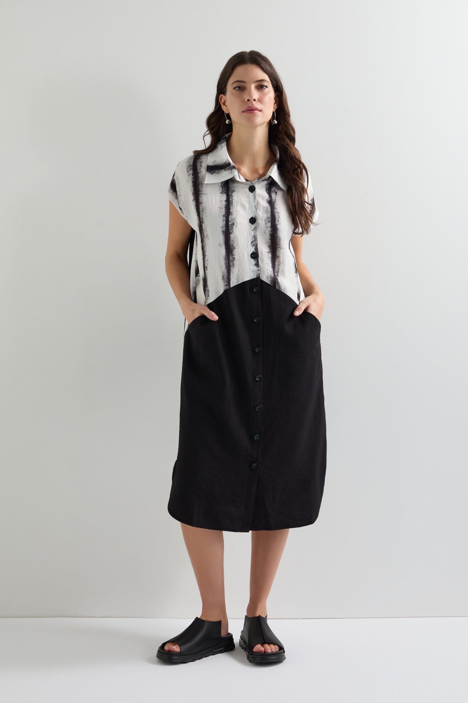 A woman models the Cap Sleeve Button Through Easy Dress, featuring black and white stripes, cap sleeves, and pockets; she pairs it with black sandals against a plain background.