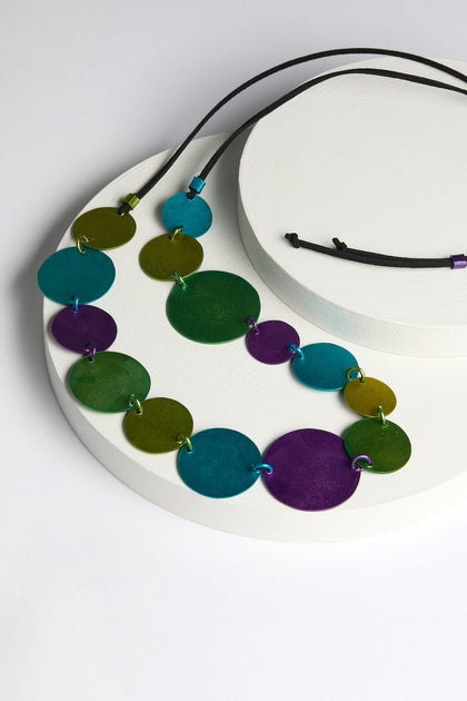 The Colour Pop Circles Necklace is a colorful, handcrafted piece featuring circular green, blue, and purple aluminum discs, elegantly arranged on a white round display. It is strung on a sleek black cord.