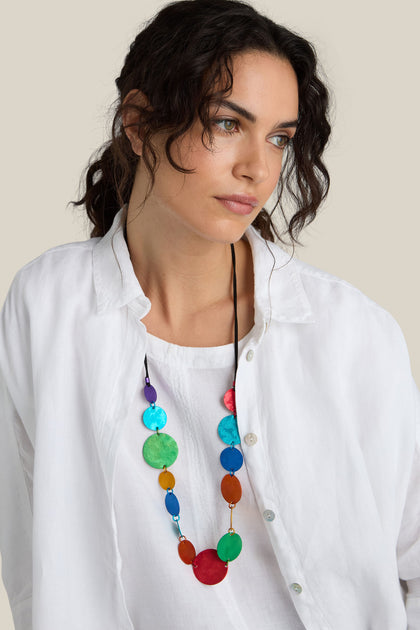 A woman with wavy dark hair wears a white shirt and the Colour Pop Circles Necklace, handcrafted by Oana Millet from aluminum discs, looking to the side with a neutral expression.