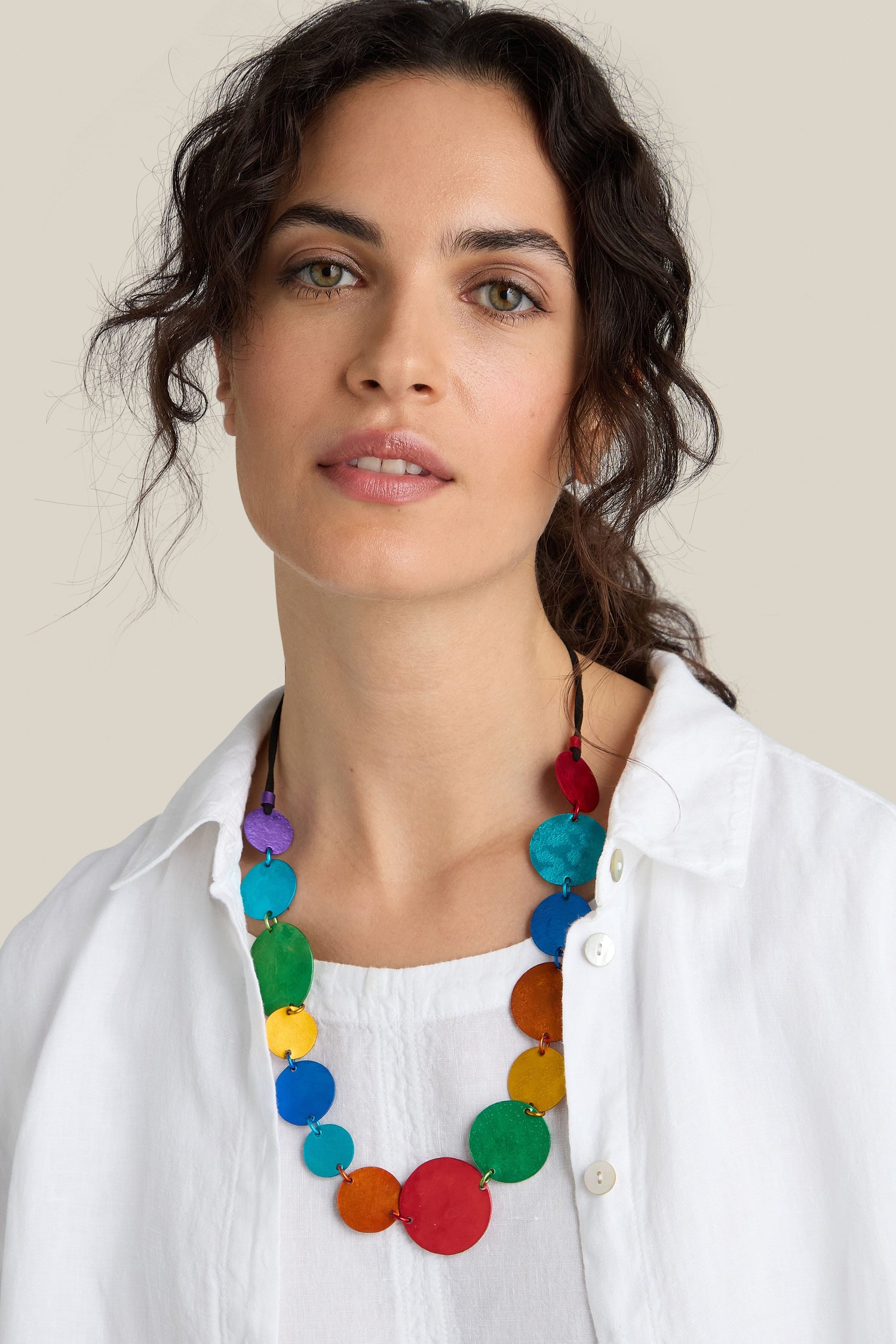 A person with wavy, dark hair wearing a white shirt and a colorful Colour Pop Circles Necklace made of handcrafted aluminium discs looks directly at the camera against a plain background.