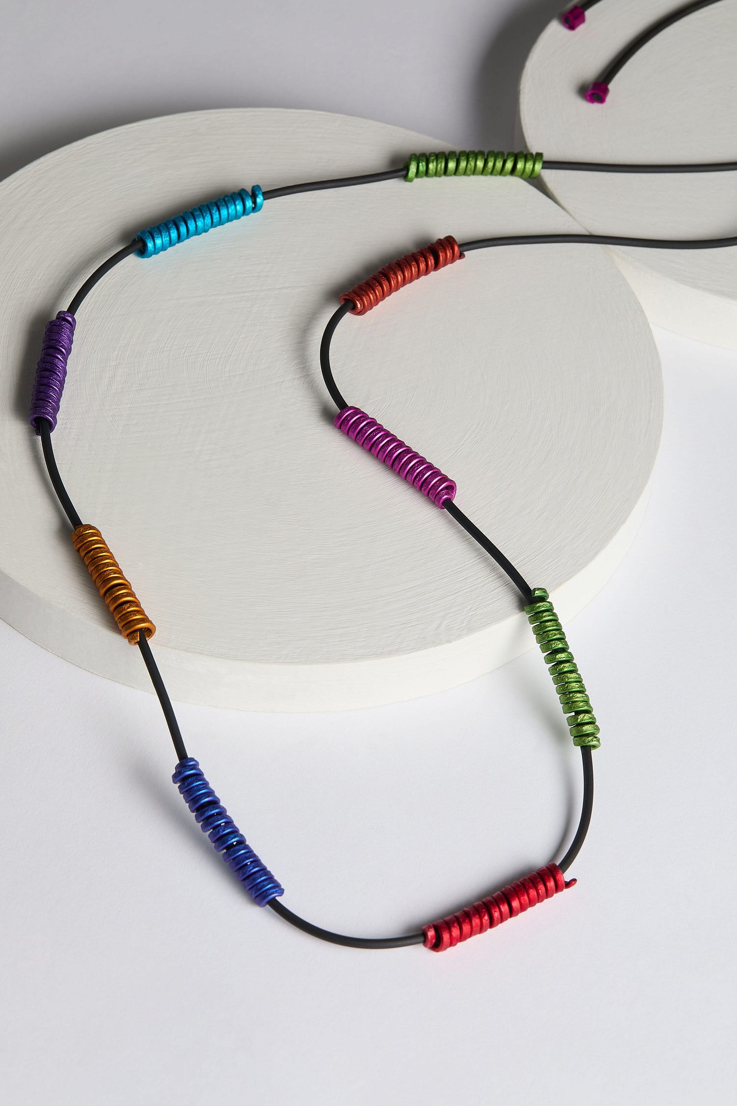 The Rainbow Spirals Necklace by Oana Millet features black cords threaded with colorful coiled metal beads, displayed on a round, white platform.