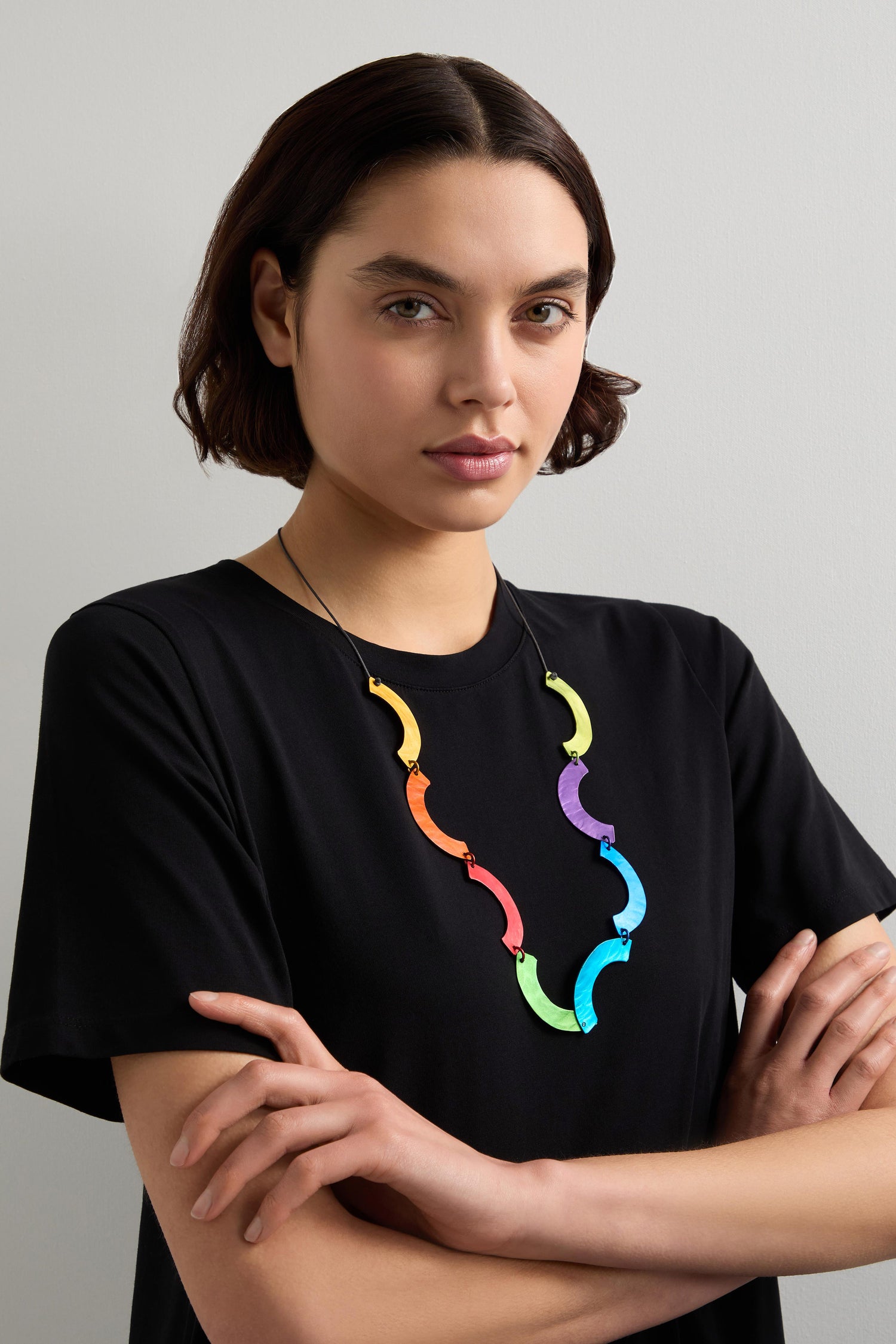 A person in a black shirt and an Aluminium Rainbow Curl Necklace stands with arms crossed against a neutral background.