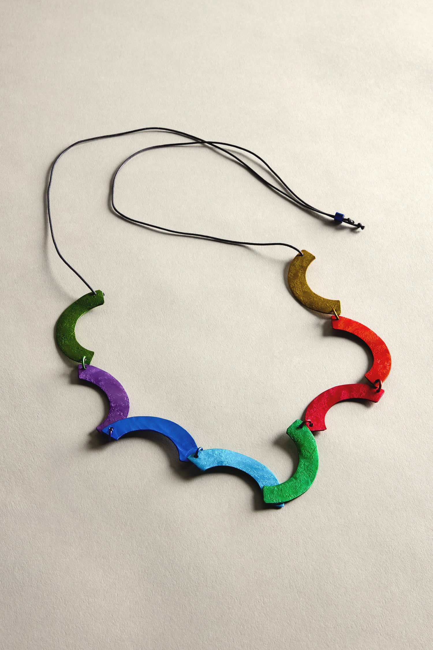 The Aluminium Rainbow Curl Necklace features a lightweight design with vibrant, crescent-shaped aluminum segments in green, purple, blue, turquoise, red, and orange on a black cord against a neutral background.