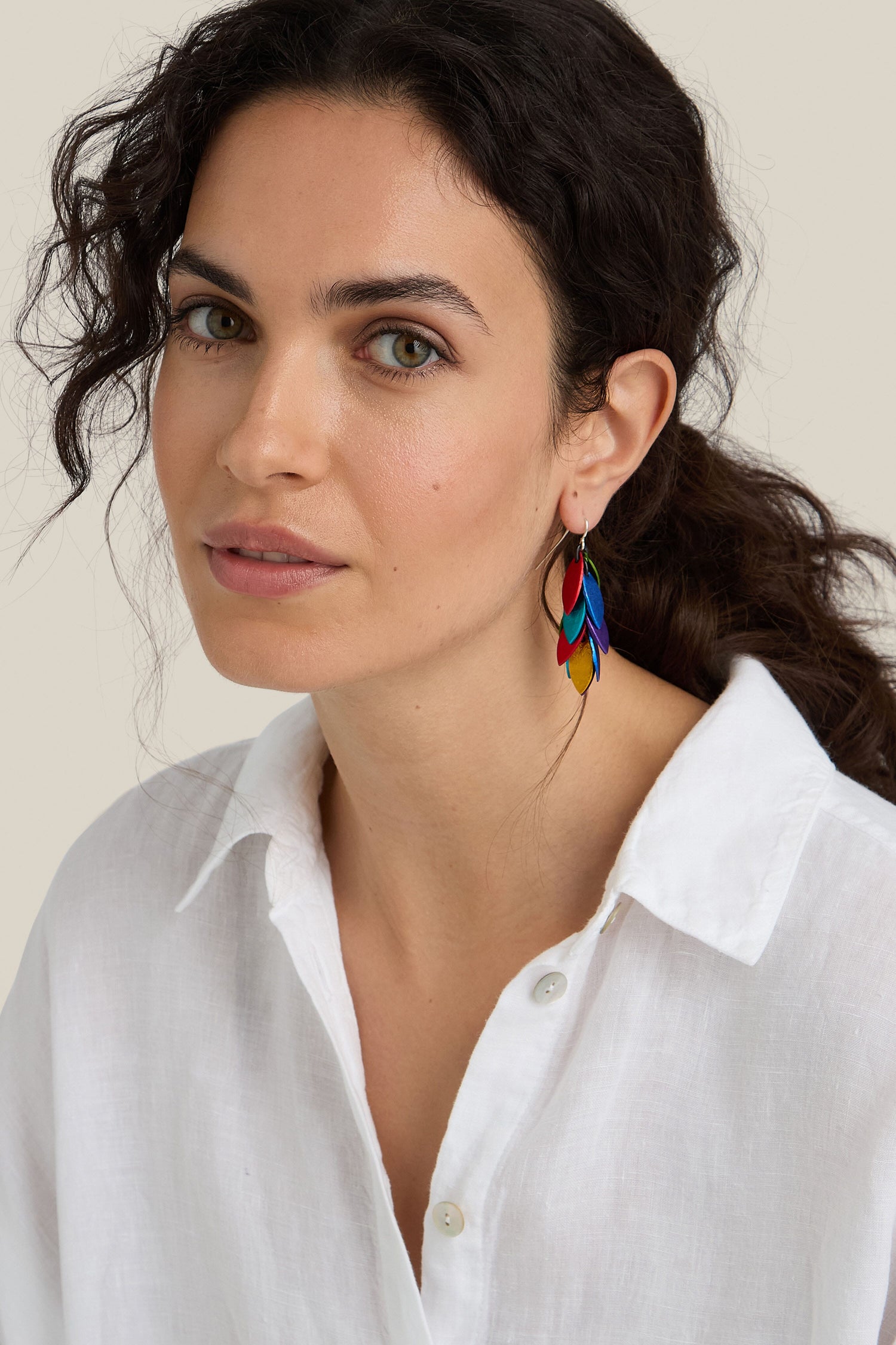 A woman with wavy dark hair wearing a white button-up shirt and the unique, colorful Scattered Leaves Earrings from a Spanish design studio gazes forward with a neutral expression against a plain background.