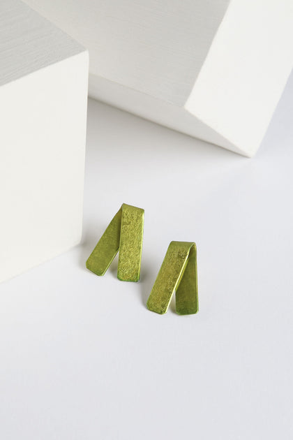 A pair of Origami Earrings in vibrant green metallic, featuring a folded rectangular design, placed on a white surface near two white geometric shapes, crafted by a renowned Spanish design studio.