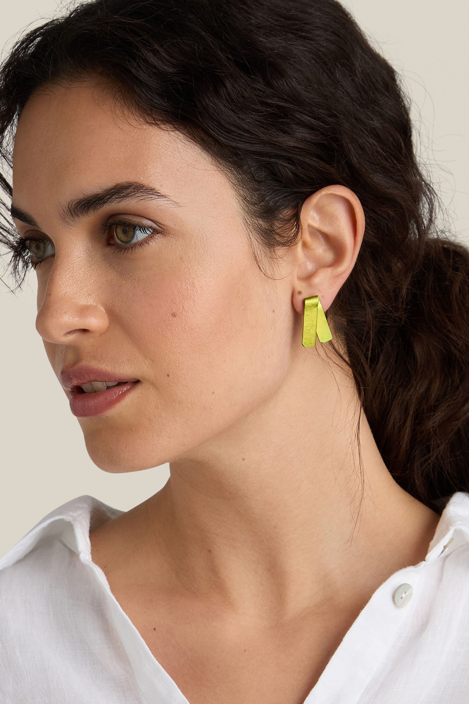 A woman with long dark hair tied back is wearing a white shirt and vibrant handmade Origami Earrings from a Spanish design studio, looking to the side.