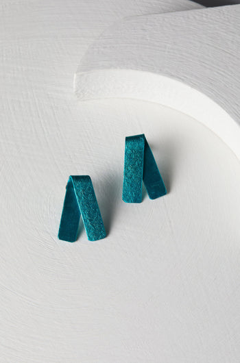 A pair of Origami Earrings in vibrant teal, crafted from folded fabric into rectangular shapes, is placed on a textured white circular surface, showcasing the exquisite craftsmanship of a Spanish design studio.