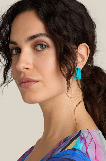 A woman with wavy dark hair wearing bright blue, handmade aluminium Origami Earrings and a colorful top looks into the camera against a neutral background.