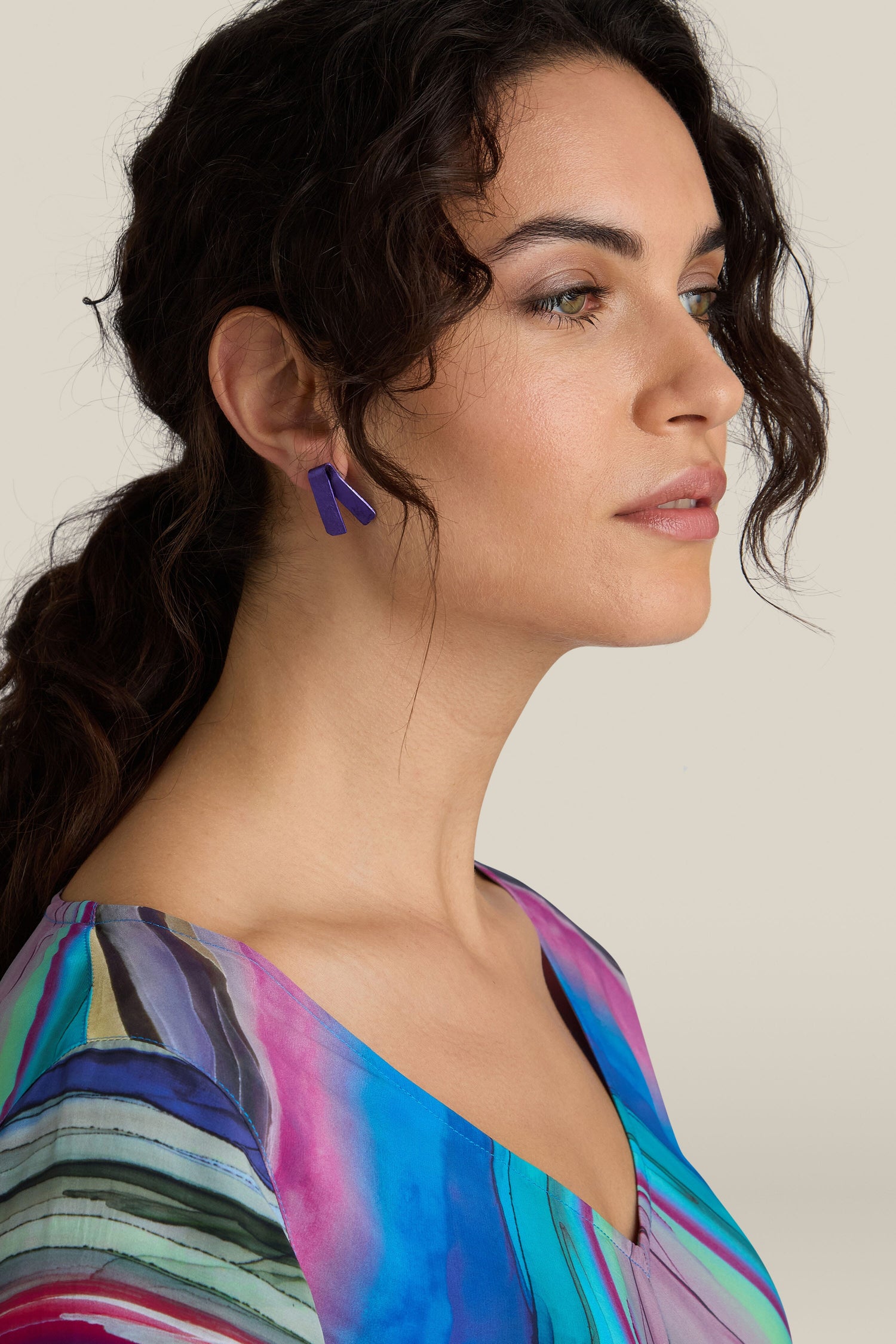 A woman with curly hair looks to the side, wearing a multicolored top and vibrant Origami Earrings from a Spanish design studio, set against a neutral background.