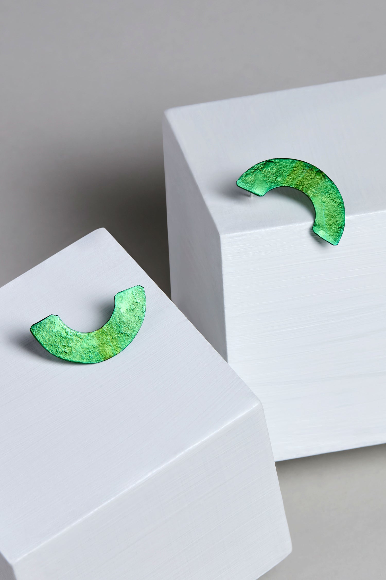 A pair of green, semi-circular Aluminium Crescent Earrings, with textured surfaces and marked OAE399, rests elegantly on white geometric blocks.