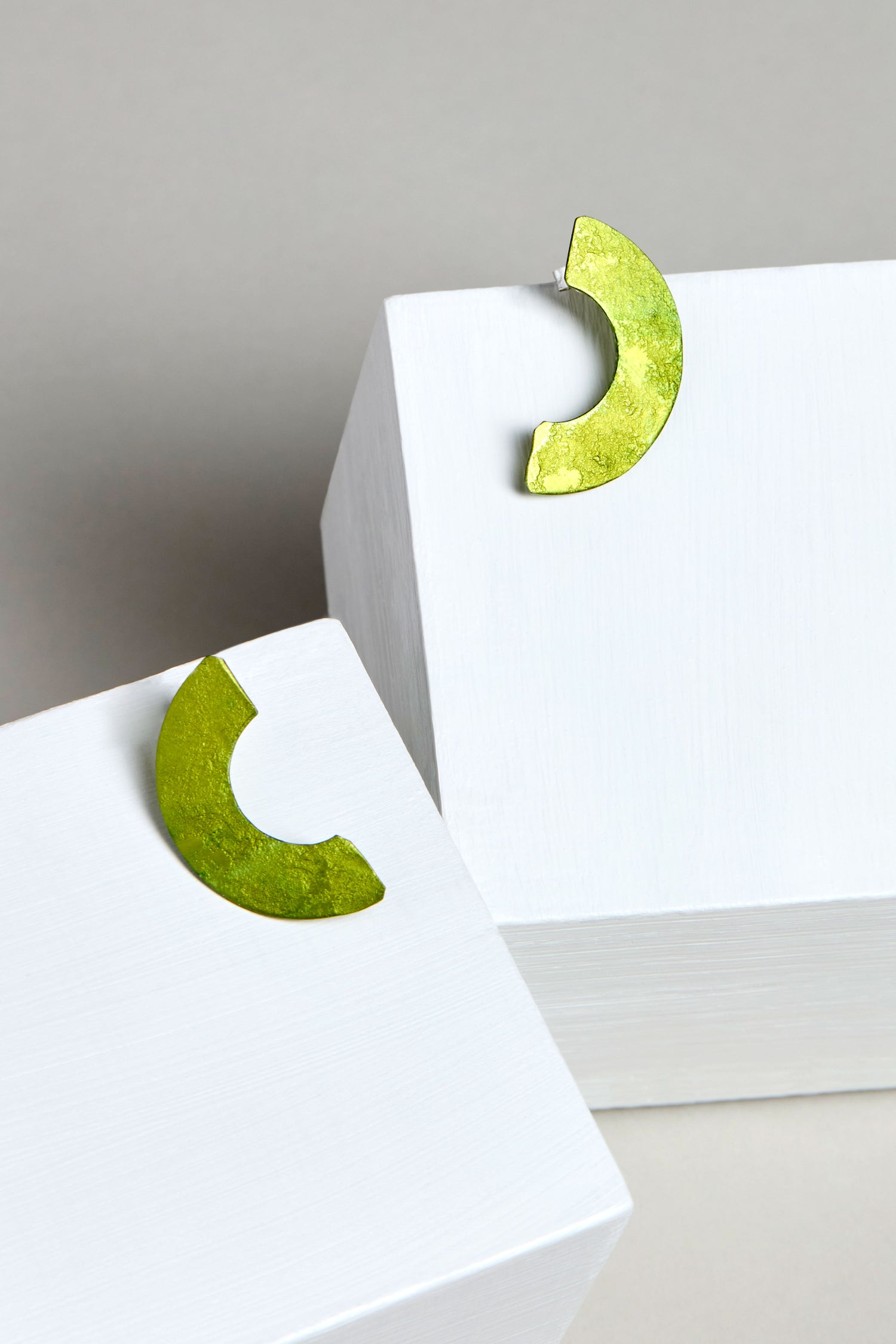 Two Aluminium Crescent Earrings in green, code OAE399, sit on white rectangular blocks against a gray background.