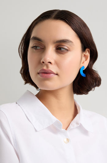 A person with short brown hair wears a white shirt and standout accessories, including blue Aluminium Crescent Earrings, looking to the side.