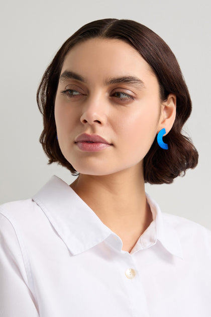 A person with short brown hair wears a white shirt and standout accessories, including blue Aluminium Crescent Earrings, looking to the side.