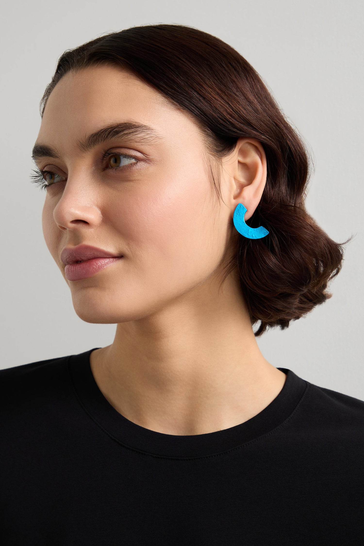 A person with short brown hair, wearing a black shirt and gaze directed to the side, is adorned with elegant blue Millet Aluminium Crescent Earrings against a neutral backdrop.