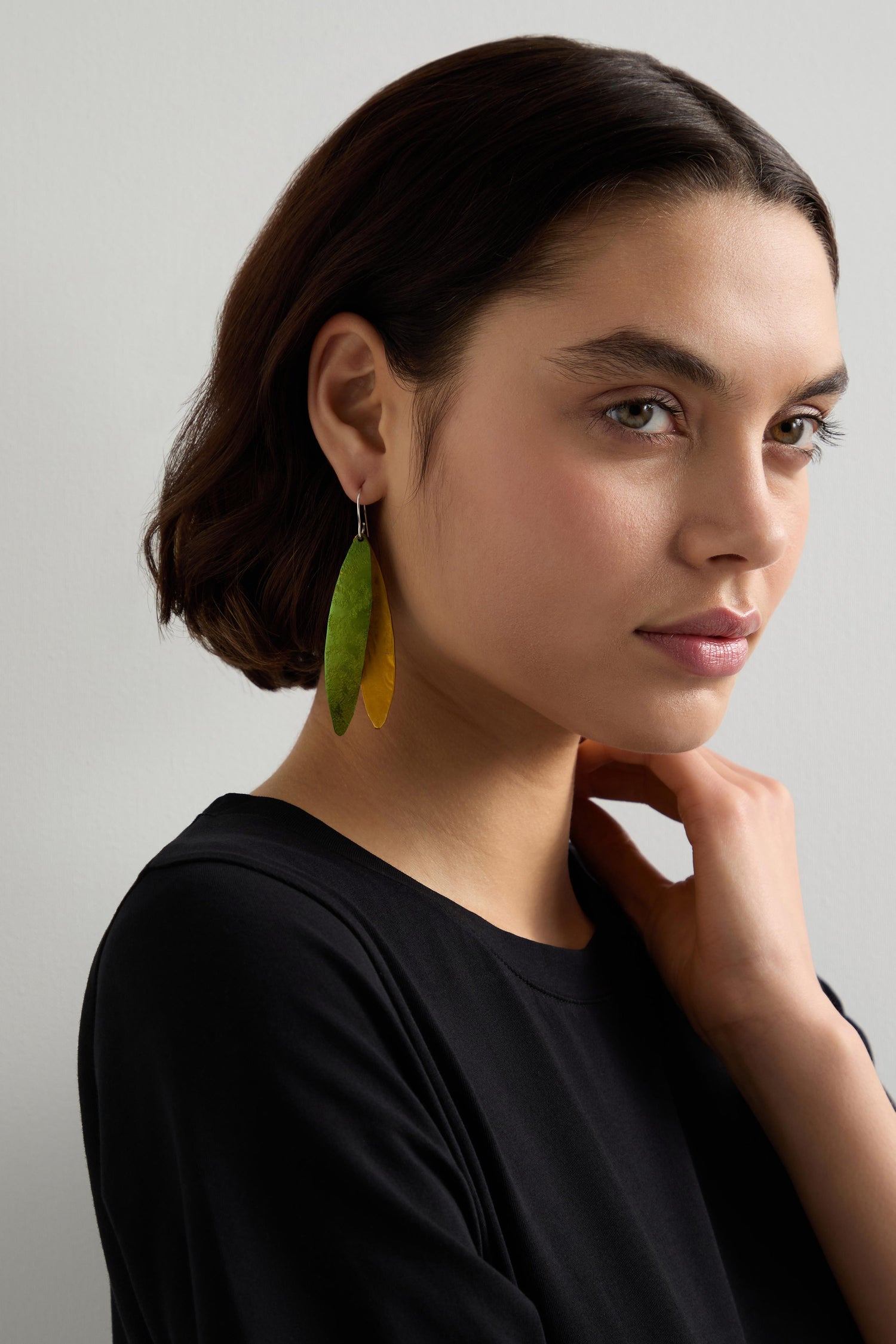 A woman with short brown hair in a black top wears large, yellow-green Aluminium Leaves Earrings by Oana Millet, posing against a light gray background and highlighting the nature-inspired design of this statement piece.