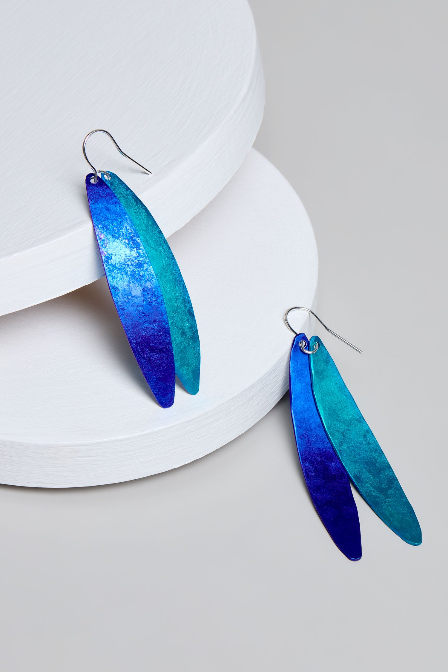 The Aluminium Leaves Earrings feature elongated, leaf-shaped designs in captivating blue and teal hues, elegantly displayed on round white platforms against a sophisticated gray background.