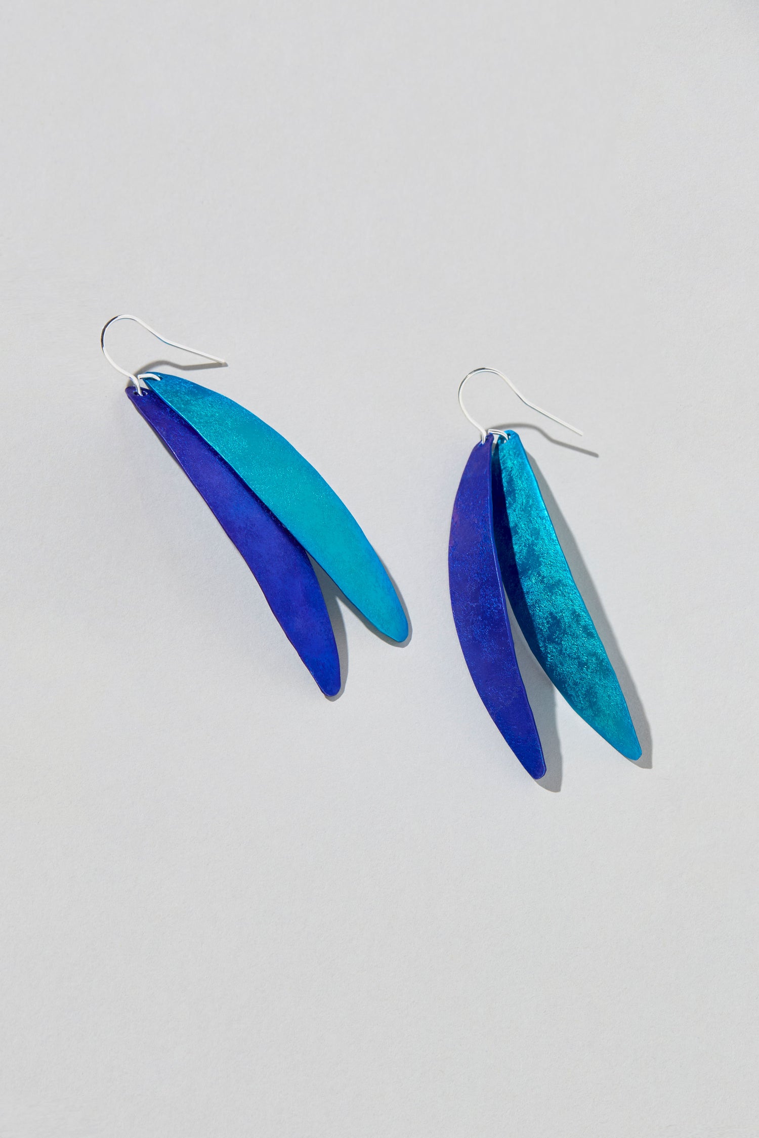 The Aluminium Leaves Earrings showcase elongated, overlapping blue leaves hanging from silver hooks, embodying serene elegance. They are ideal for adding a touch of nature-inspired sophistication to any outfit.