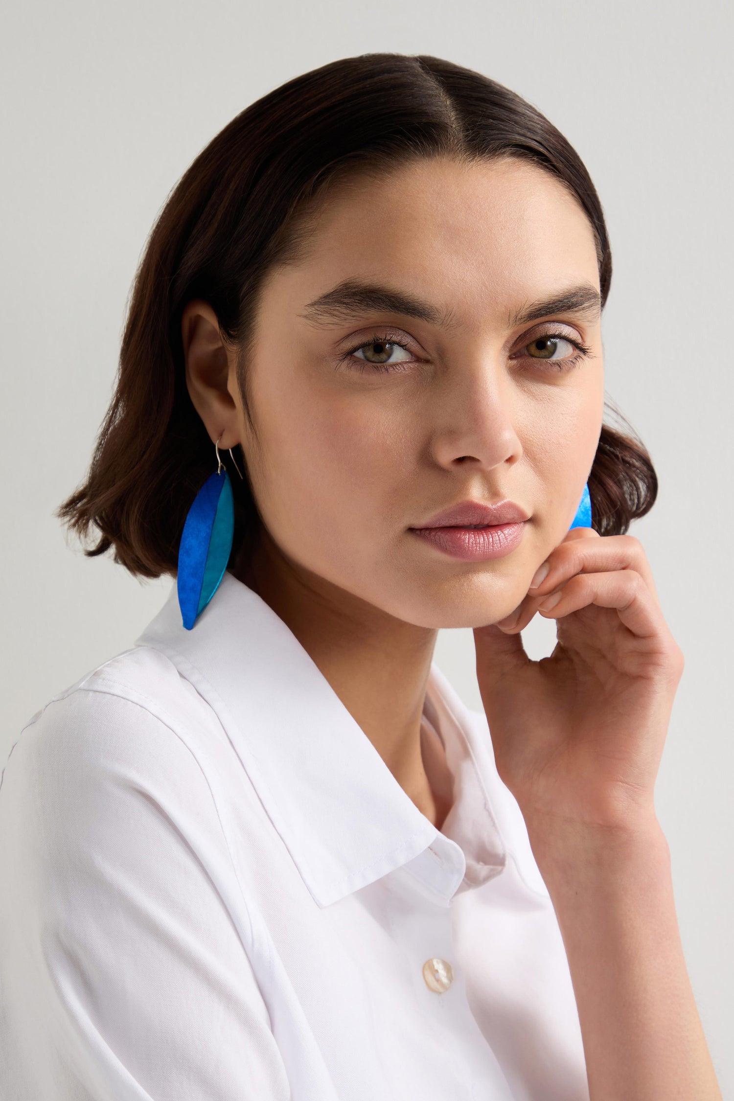 A person with short dark hair and a white shirt touches their face, wearing handcrafted Earrings in the Aluminum Leaves design, as they look at the camera.