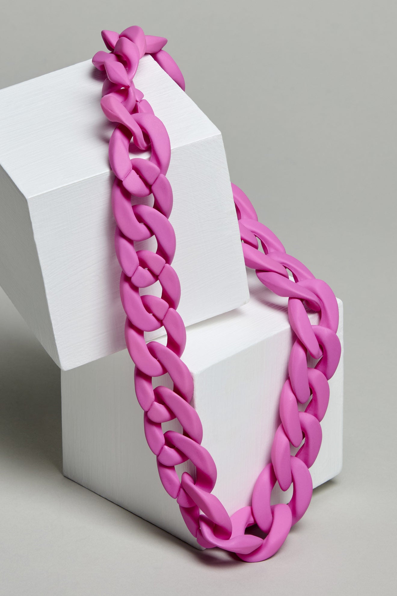 The Colour Pop Link Necklace, a bold and chunky pink chain, drapes elegantly over two white geometric blocks on a gray background.