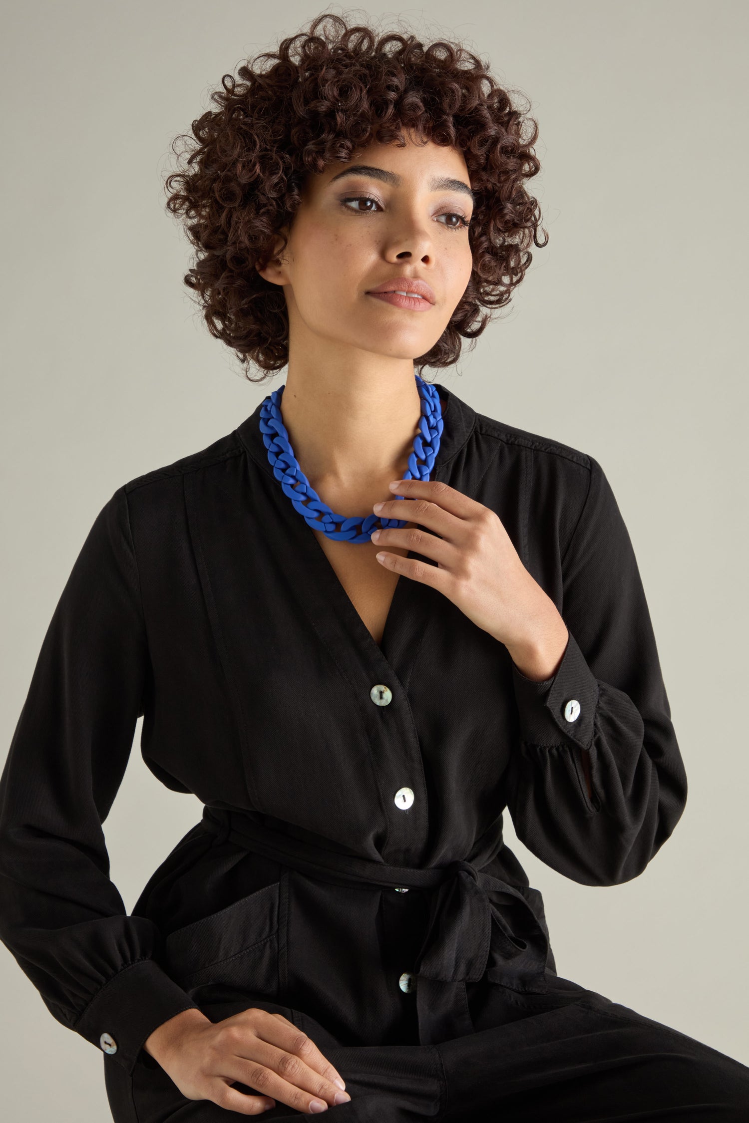 A woman with curly hair poses against a neutral background, wearing a black outfit and the Colour Pop Link Necklace.