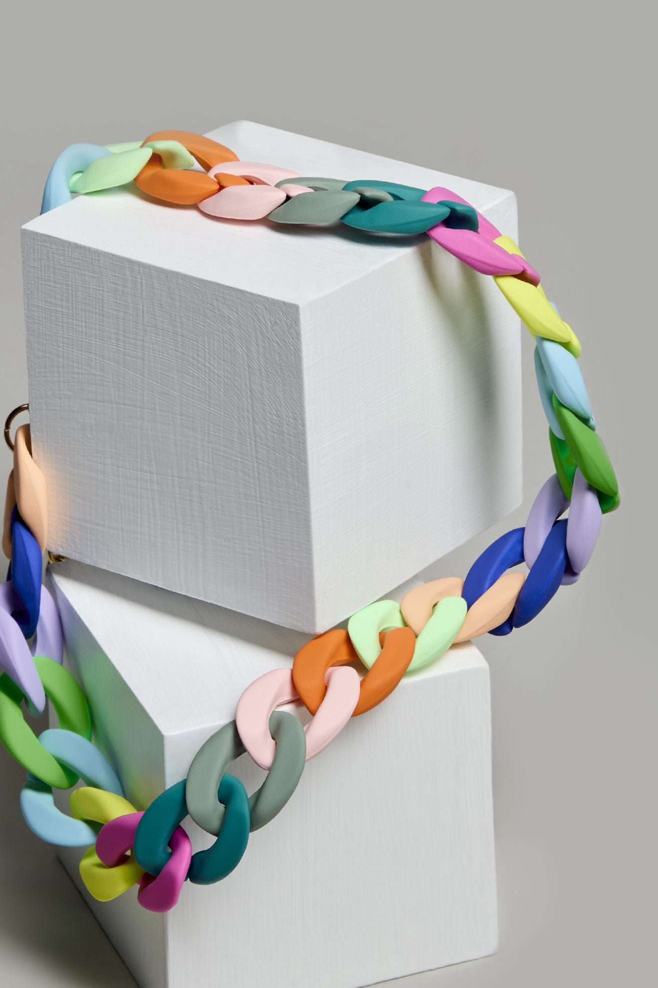 The Colour Pop Link Necklace, with its vibrant interlocking design, is elegantly draped over two stacked white cubes against a gray background to create a stunning statement piece.