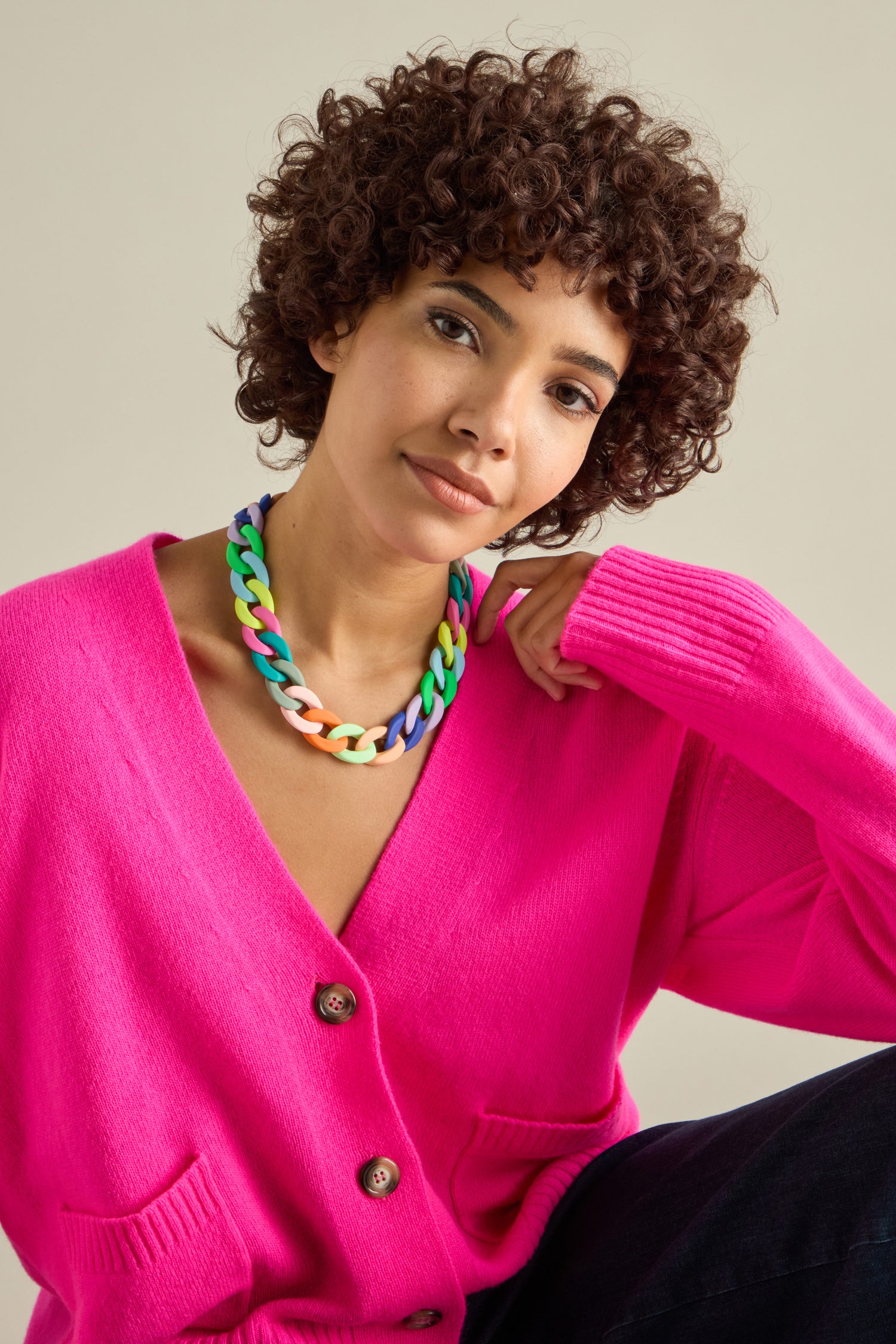 A person with curly hair is wearing a bright pink cardigan and the Colour Pop Link Necklace, sitting against a plain background with one arm resting on their knee.