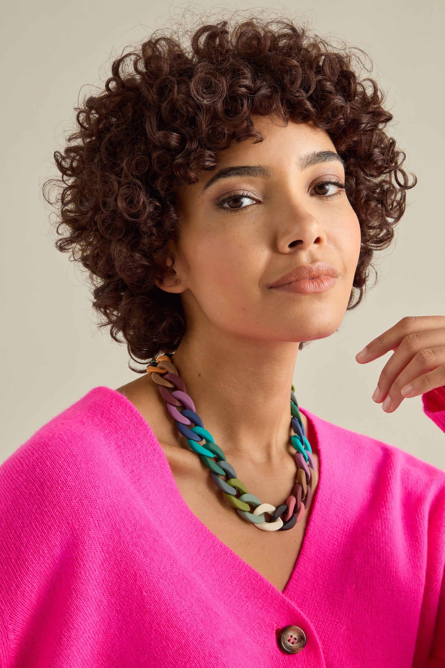 A person wearing a bright pink cardigan and a bold statement piece—a Colour Pop Link Necklace—looks directly at the camera.