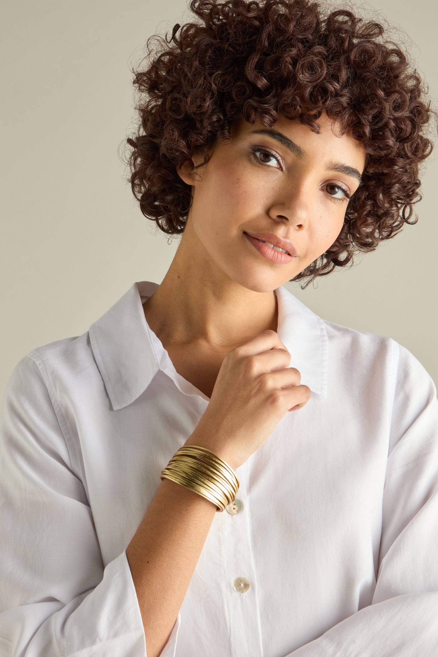 A person with curly hair wearing a white shirt and a stack of Woven Metallic Bracelets smiles slightly, looking at the camera.