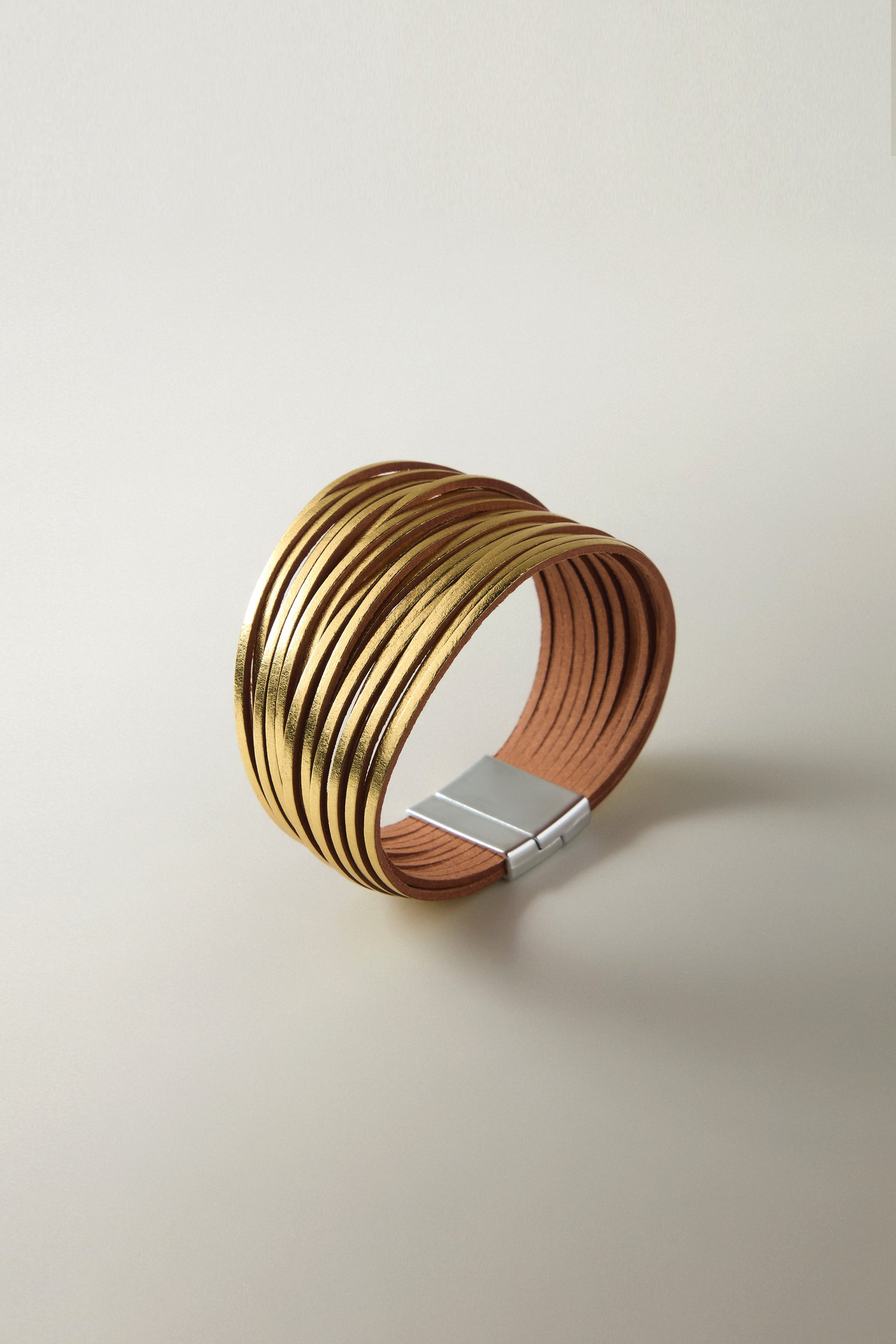 The Woven Metallic Bracelet features a gold and brown metallic finish with multiple stacked bands, adorned with leatherette threads and finished with a silver clasp, showcased on a neutral background.